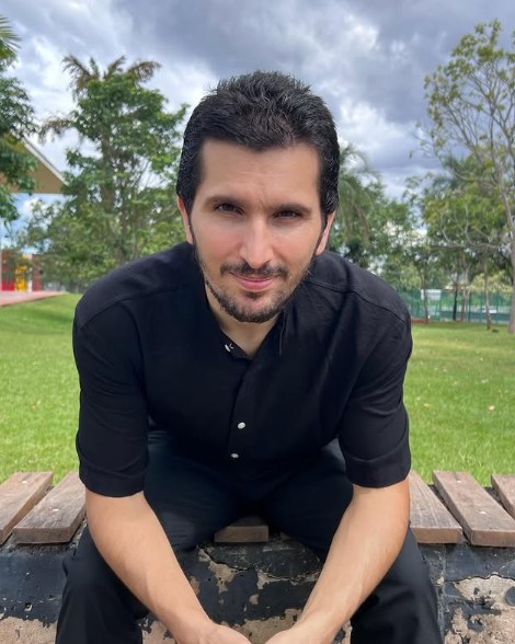 Rafael Bianco sentado em um banco de praça, de camisa preta, olhando para a câmera